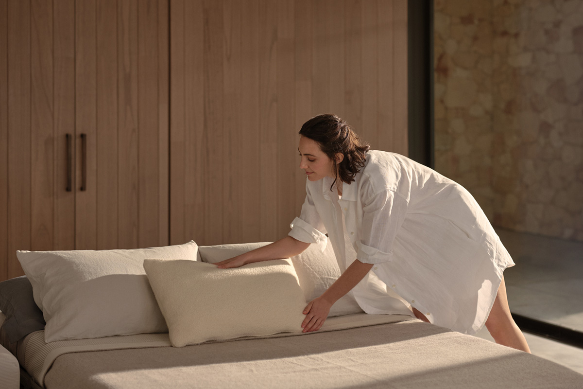 Woman arranging pillows on a neatly made bed in a serene, light-filled room with wooden accents.