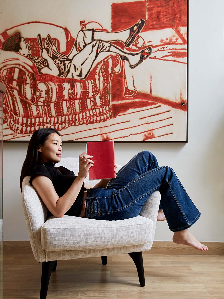 Woman sitting on a cream armchair reading a red book with artwork in the background.